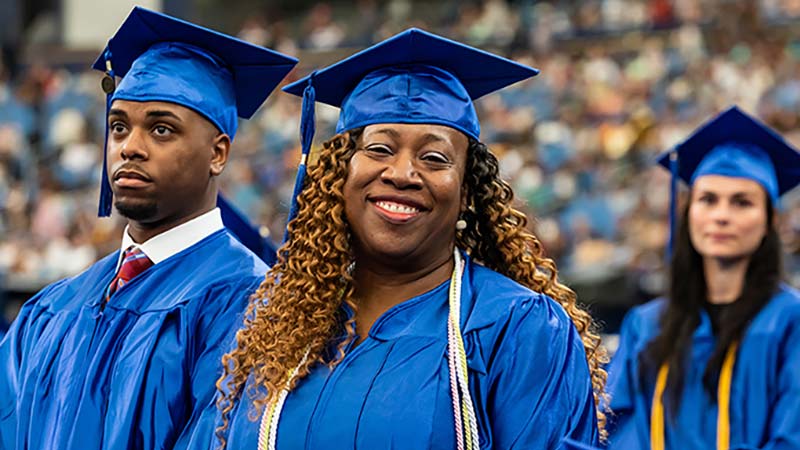 Wearing their blue caps and gowns, a female graduate stands and smiles for the camera at an SPC graduation ceremony with a male ad female student standing behind her.