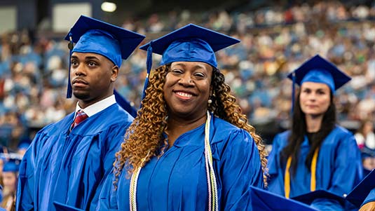 Wearing their blue caps and gowns, a female graduate stands and smiles for the camera at an SPC graduation ceremony with a male ad female student standing behind her.