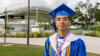 SPC graduate Dominick Dos Santos in his blue cap and gown standing in front of the The BayCare Sound in Clearwater 