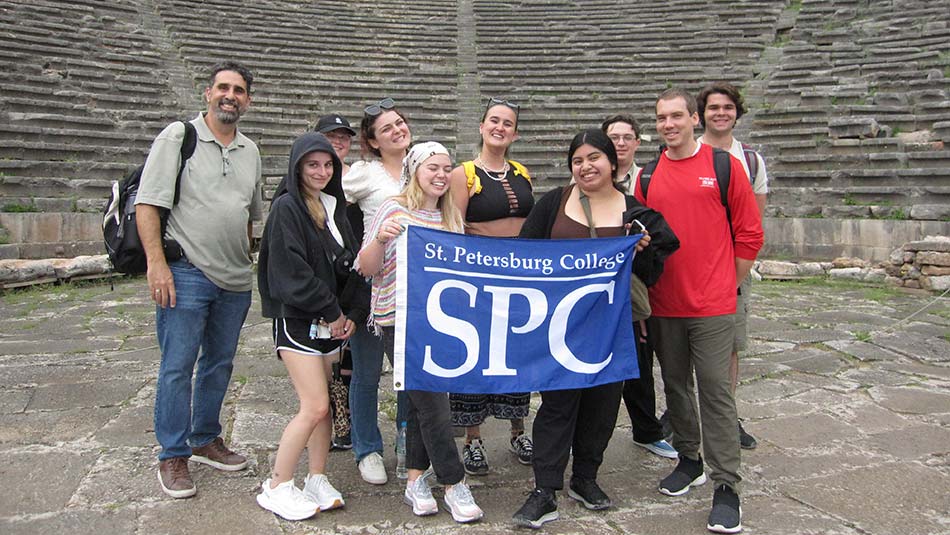 A group of students hold an SPC sign in front of a beautiful backdrop of ancient Greek ruins.