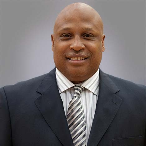 Portrait of Earnest Crumbley, over a gray background, wearing a black suit coat over a white dress shirt with gray stripes, and a black and white striped tie.