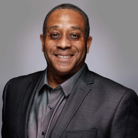 A portrait of Mark McKinney, wearing a dark gray suit over a lighter gray dress shirt. 