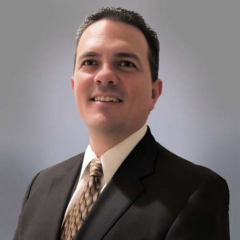 A portrait of Wayne Kruger over a gray background, wearing a black suit coat, white dress shirt and a multi-tone brown tie.