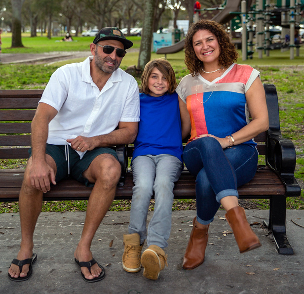 Luisa Shamas sits on a park bench with her husband and son, a children's playground in the background