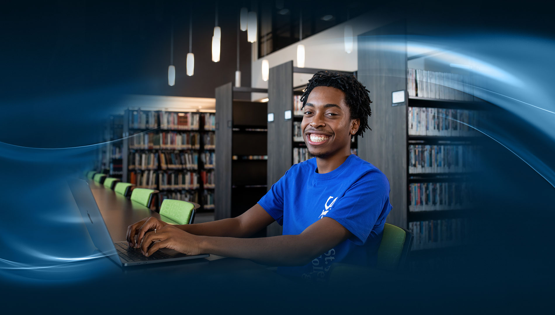 male SPC student in a blue SPC t-shirt sitting at a laptop smiling broadly
