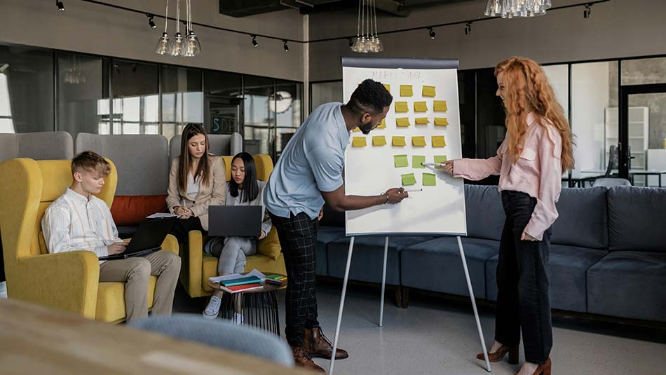 A man and woman in front of a white board with sticky notes all over it, as .3 other people, seated in chairs, work on laptops in the background.